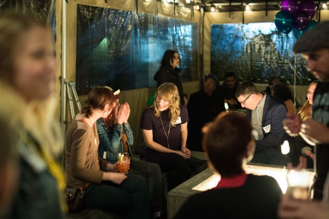 Group of attendees socializing at the Love Bomb 2017 event, seated around a lit table, with drinks in hand, capturing the community spirit and engagement supporting My Voice Music.